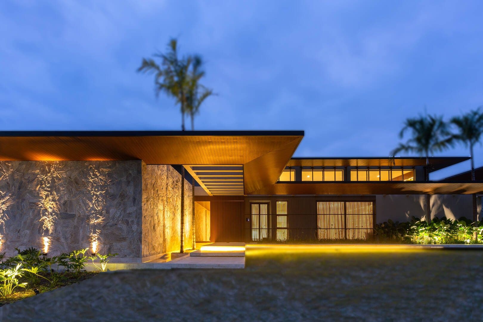 Fachada de arquitetura autoral ao crepúsculo, com madeira, pedra e iluminação quente