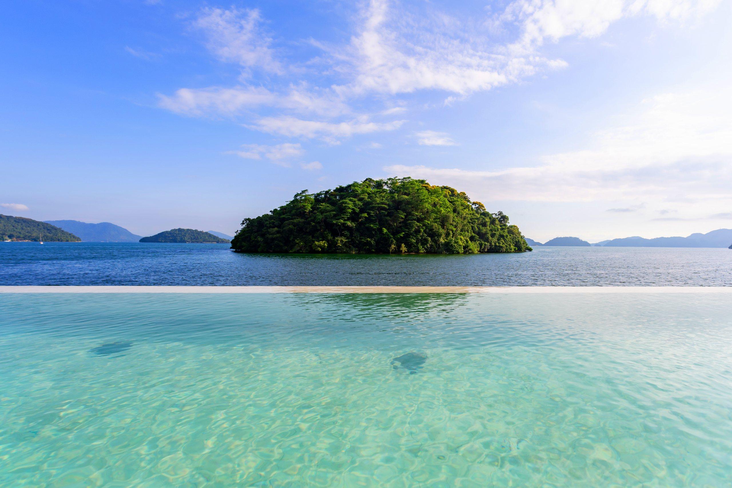 Piscina de borda infinita à beira-mar, com a água transparente se fundindo à baía e uma ilha arborizada ao fundo, em Angra dos Reis
