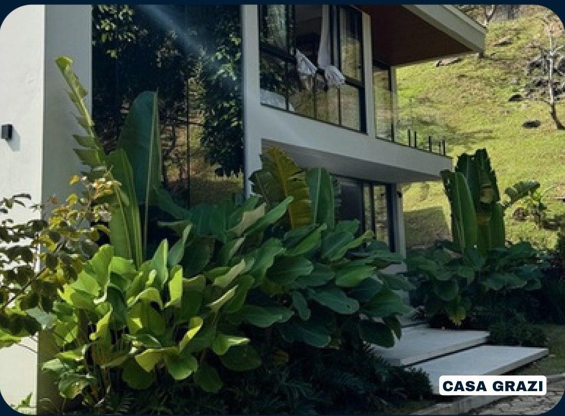 Casa Grazi entre vegetação densa, com volumes brancos e vidro voltado para o vale
