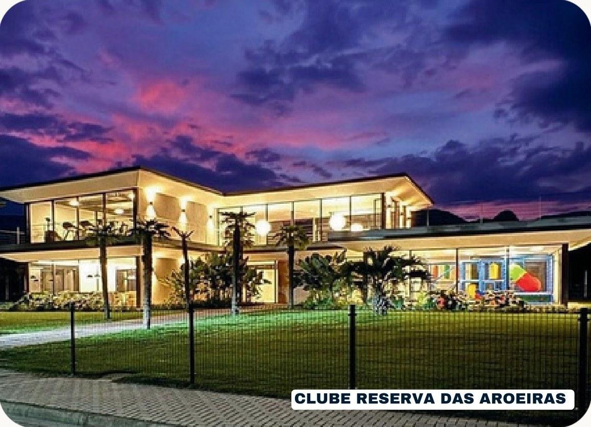 Clube Reserva das Aroeiras iluminado ao entardecer, com gramado e fachada envidraçada