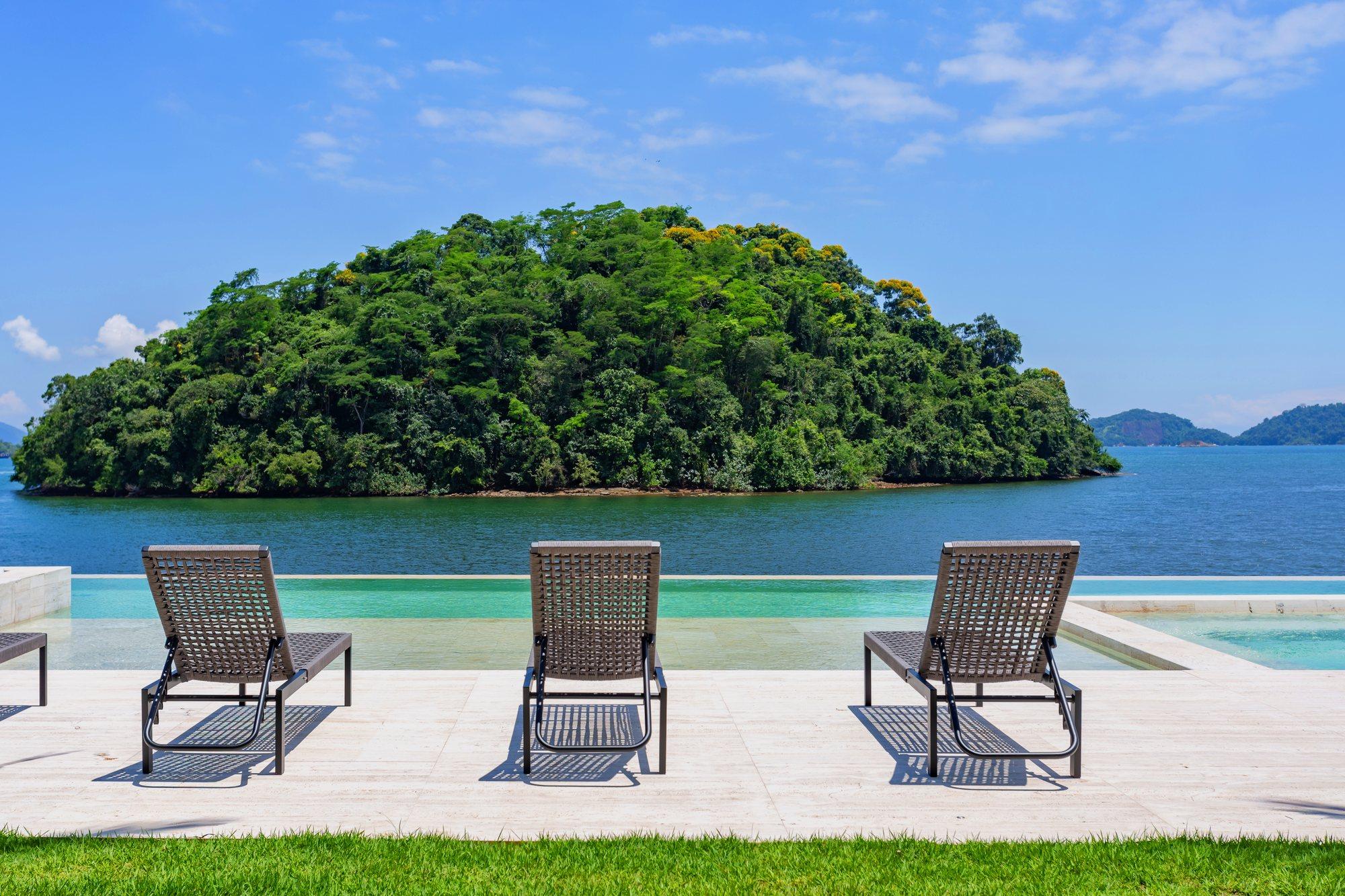Espreguiçadeiras à beira da piscina de borda infinita, com uma ilha arborizada e o mar de Angra ao fundo